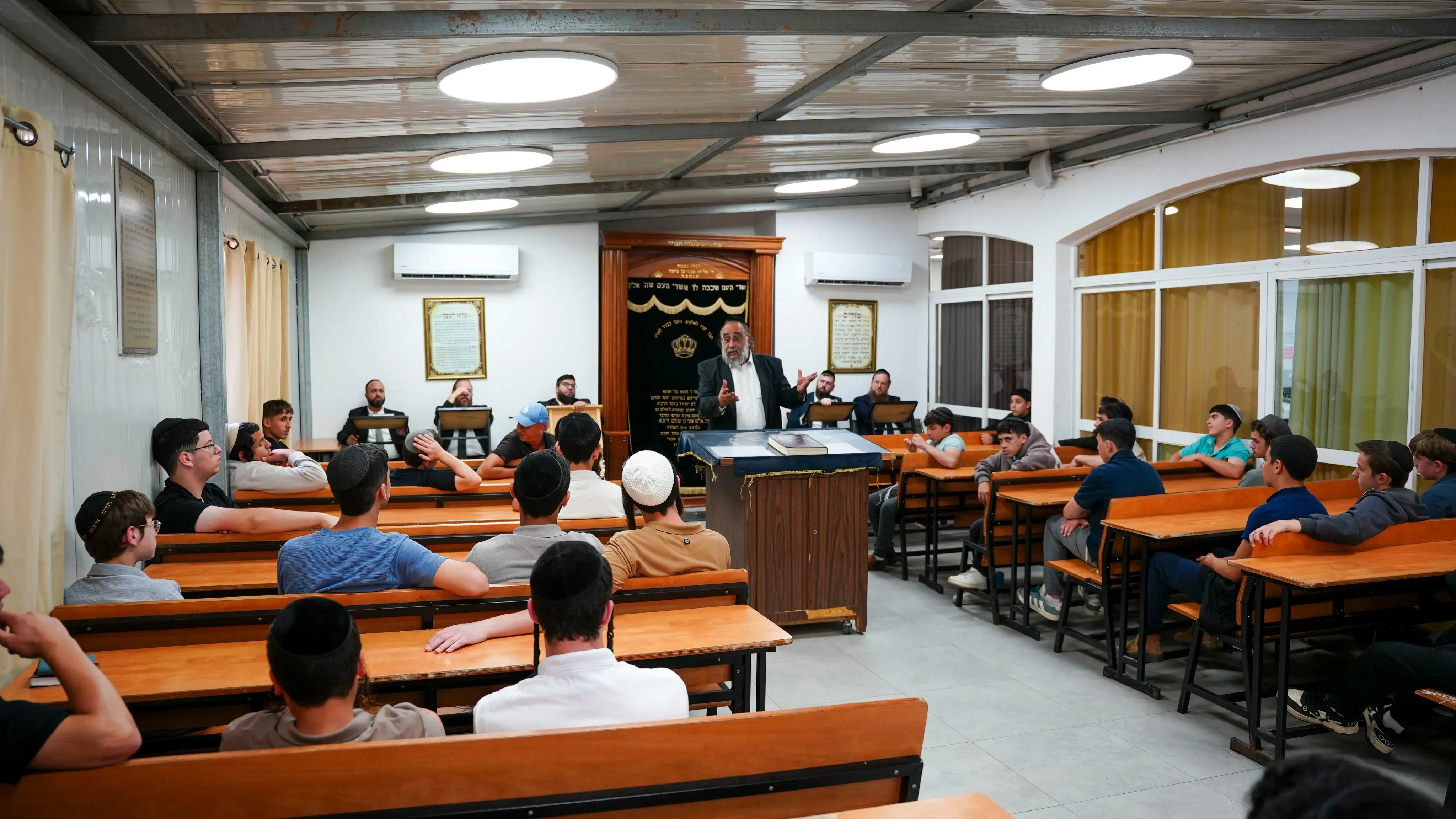 תלמידים ורבנים מתאספים באולם בית המדרש מול ארון הקודש, רב עומד ונושא דברים