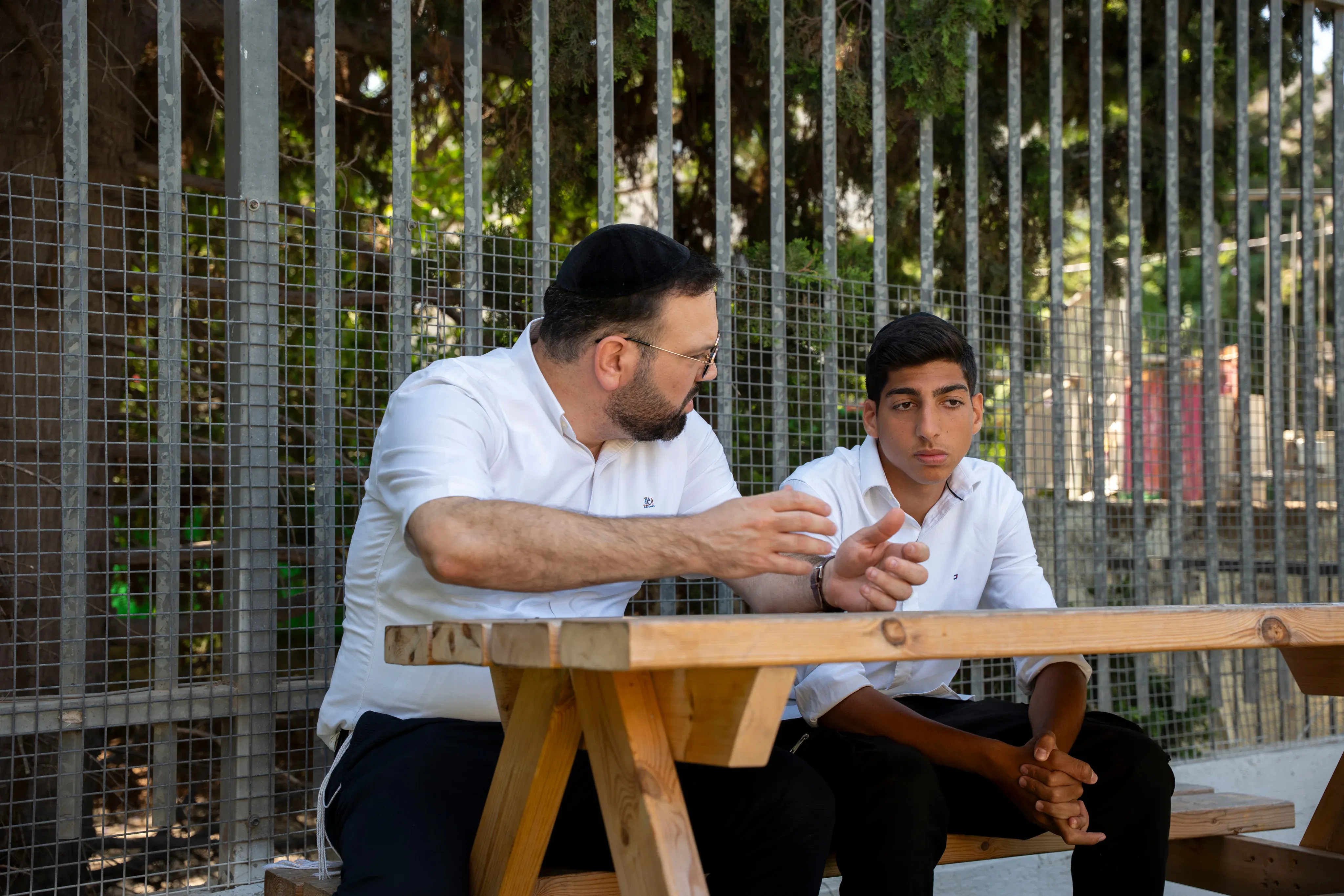 רב בשיחה אישית עם תלמיד, יושבים יחד ליד שולחן פיקניק בחוץ, גדר רשת ועצים ברקע