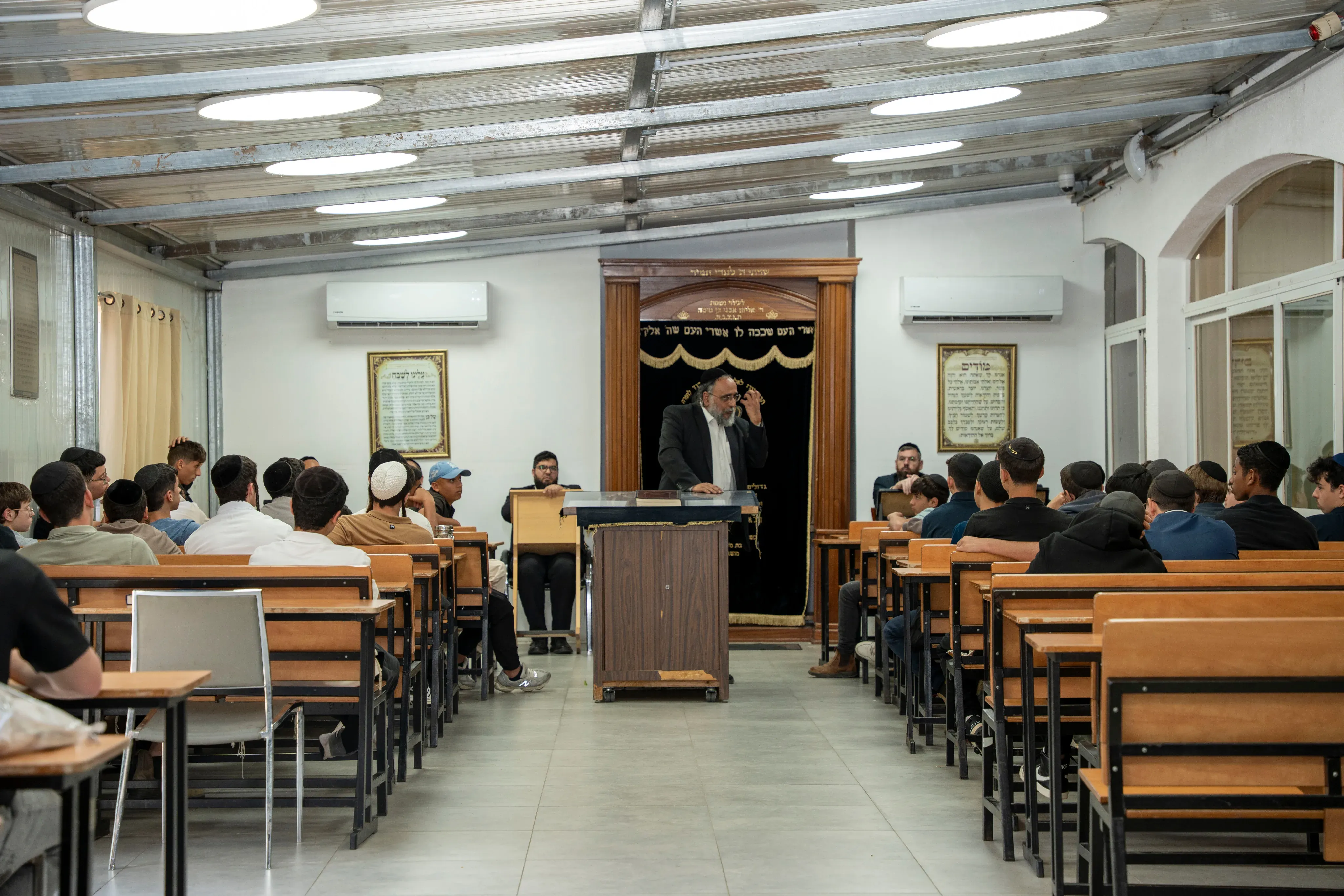 רב עומד בעמדת הדרשה מול ארון הקודש ונושא דברים לפני התלמידים היושבים באולם