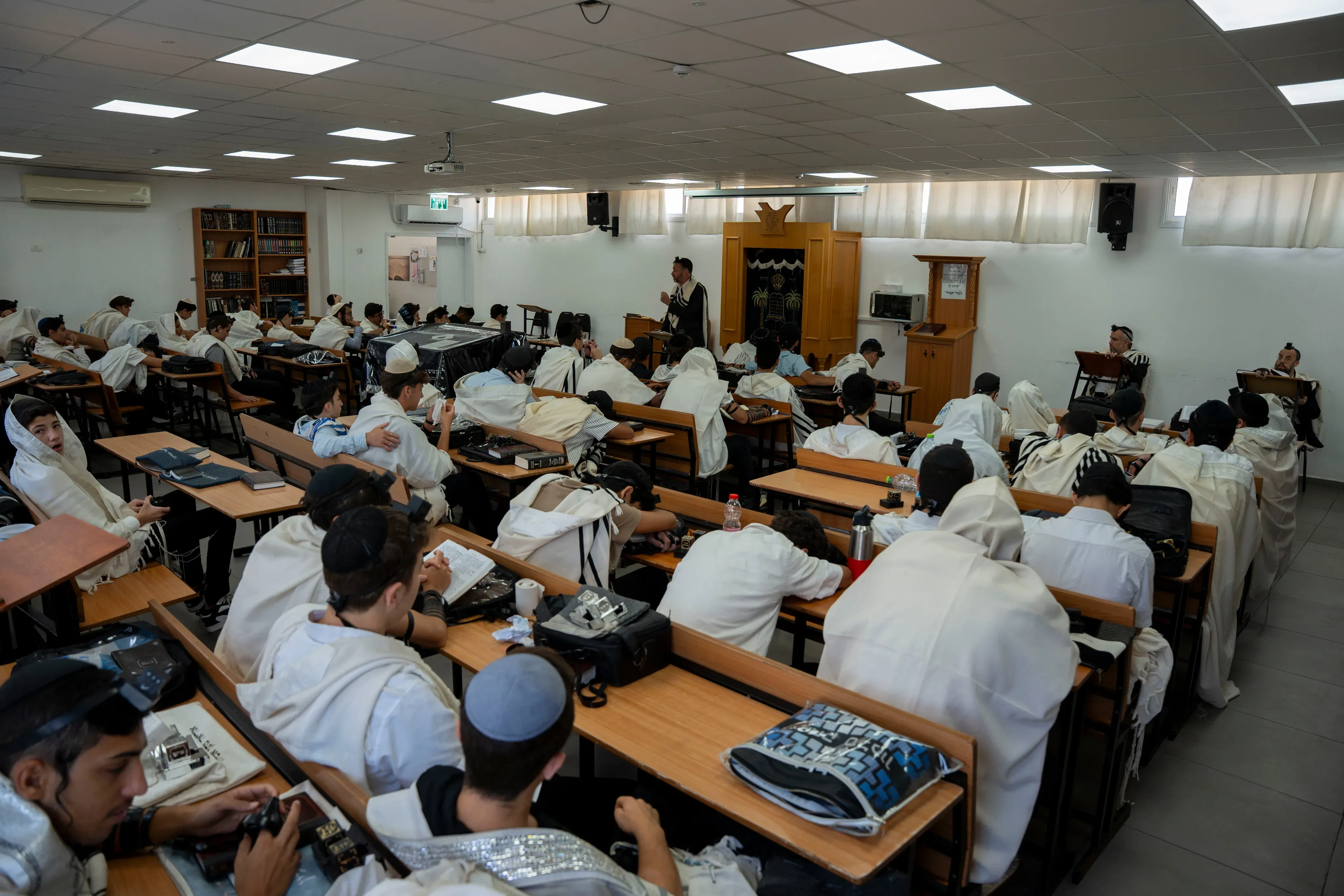 תלמידים עטופים בטלית בתפילת שחרית באולם בית המדרש, רב עומד ומתפלל בראש האולם