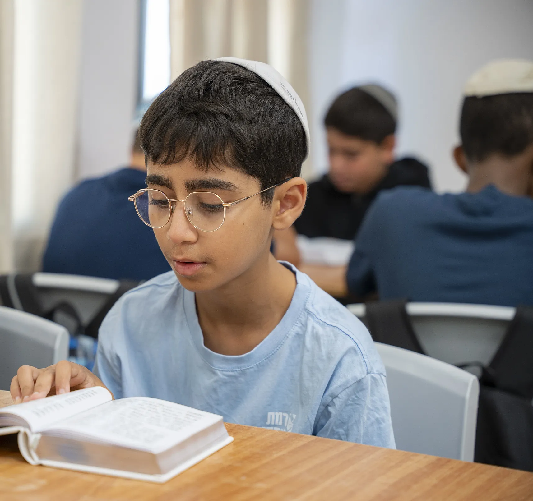 תלמיד במשקפיים קורא בקול מתוך ספר ליד שולחן בכיתה