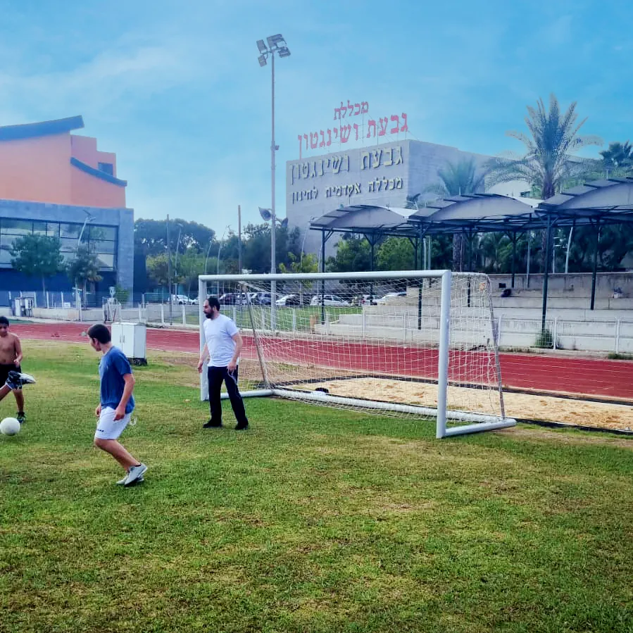 כדורגל על הדשא מול שער ומבנה הספורט - יוצאים למגרש