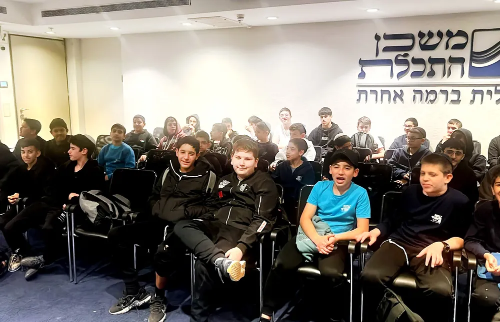 תלמידים יושבים באולם משכן התכלת מבט רחב - רמת אקדמי באר יעקב, קושרים ציצית