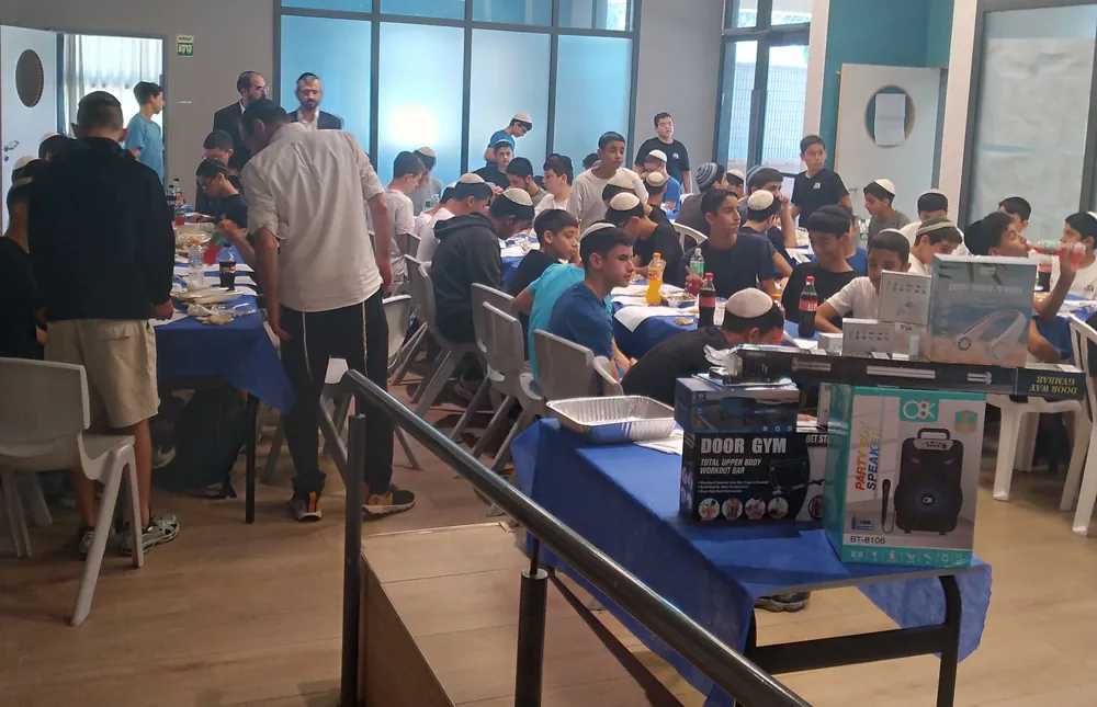 תלמידים יושבים בחדר מלא סביב שולחנות, ליד שולחן עמוס בקופסאות פרסים ומתנות