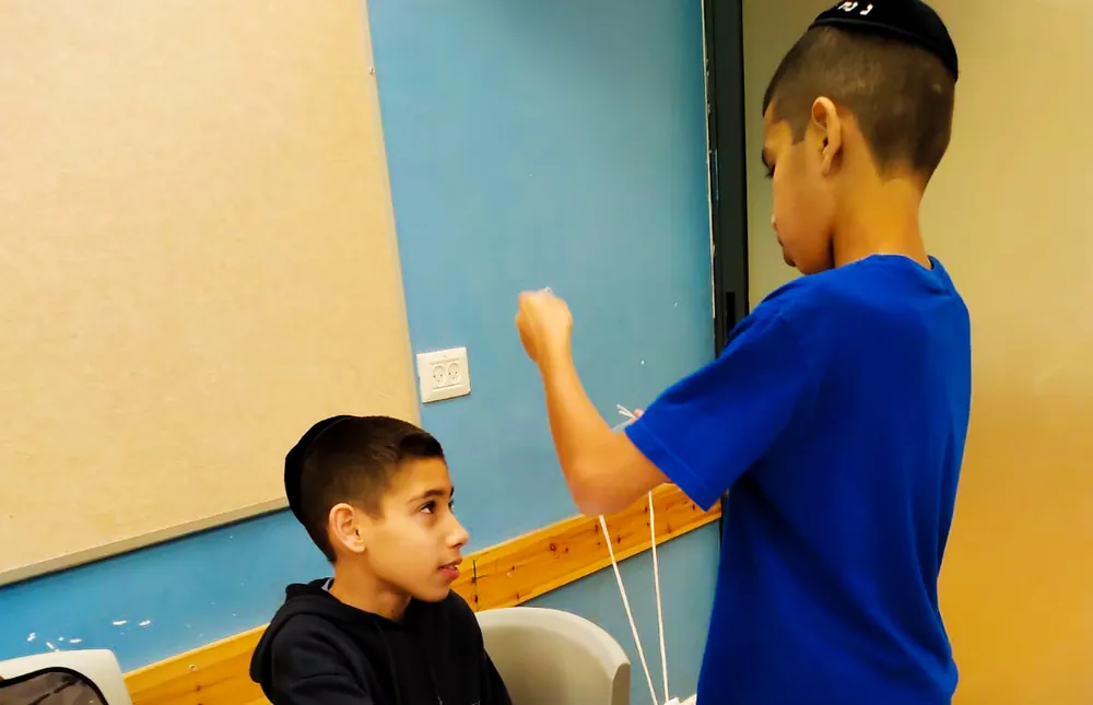 שני תלמידים מכינים חוטי ציצית ליד שולחן - לומדים לקשור ציציות