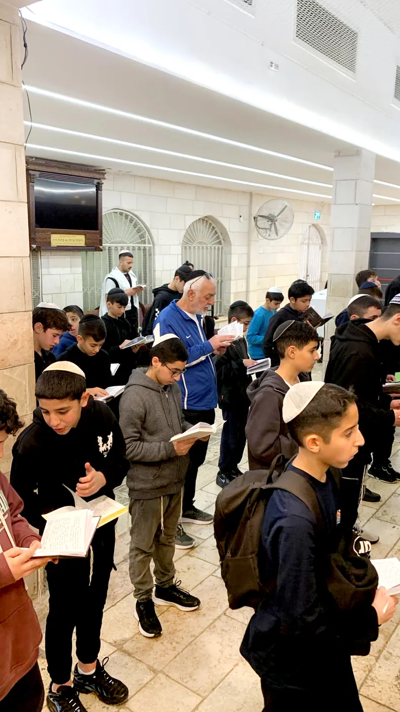 תלמידים ומבוגר קוראים בסידורים בבית כנסת - לומדים לקשור ציציות