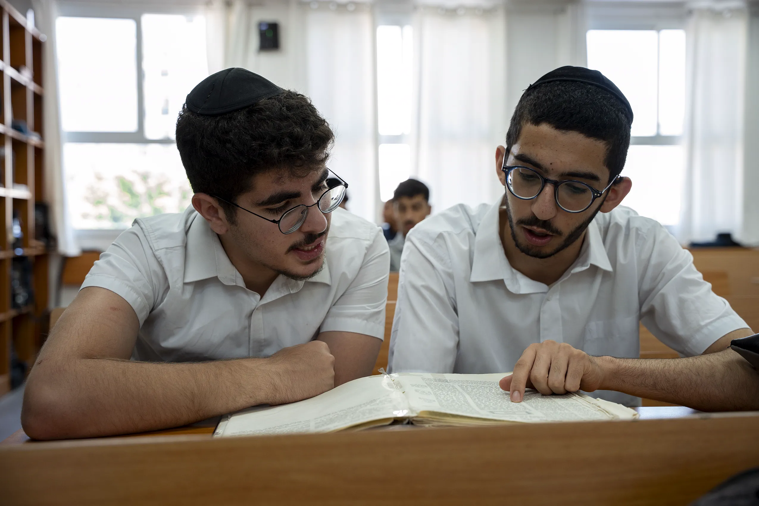 חברותא: שני תלמידים לומדים גמרא מעל ספר, מדפי ספרים ברקע
