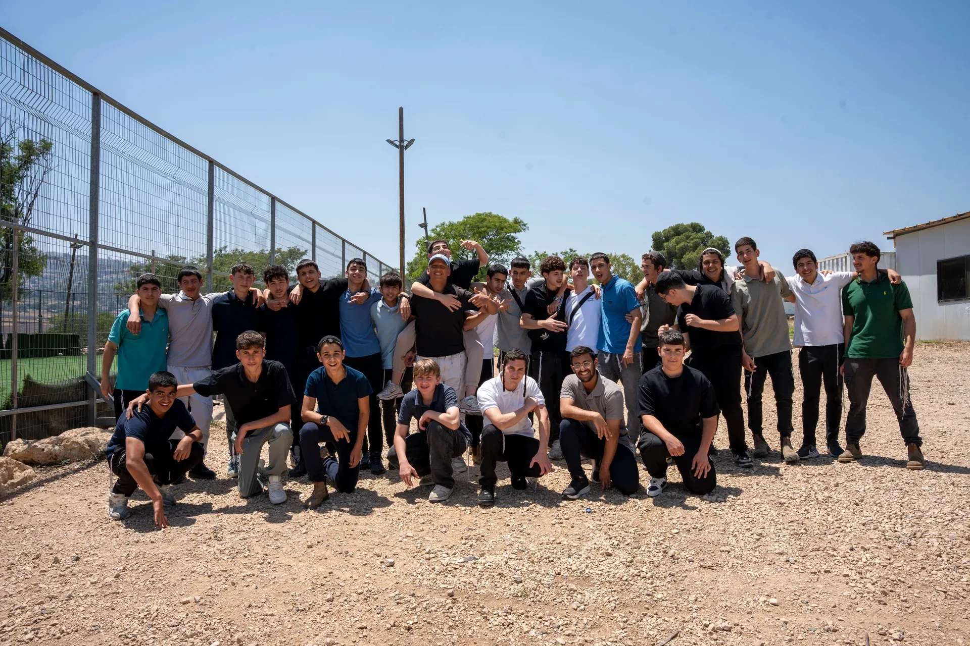קבוצת תלמידים עומדים ויושבים יחד בחוץ, במגרש מגודר