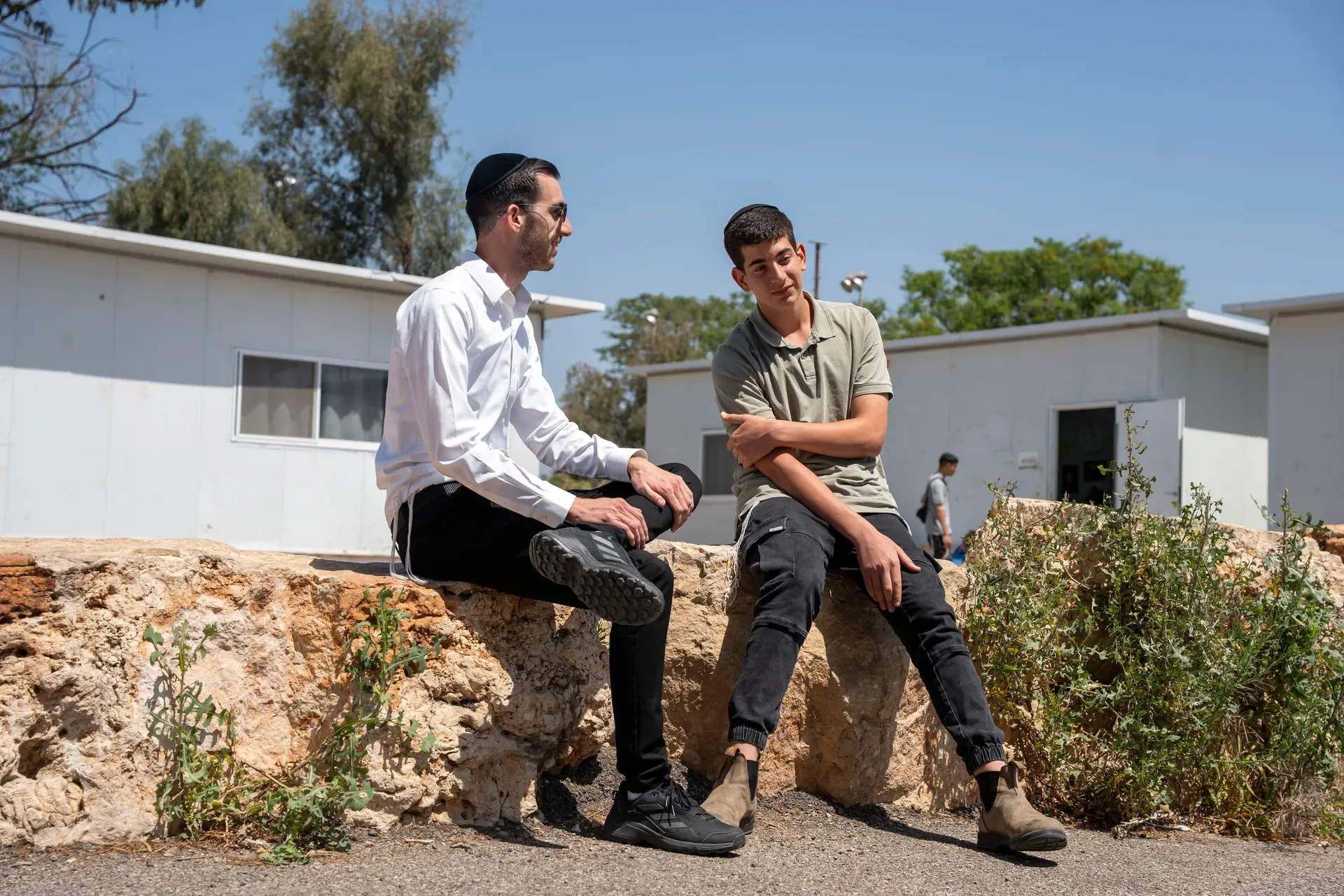 רב ותלמיד משוחחים כשהם יושבים על קיר אבן בחצר