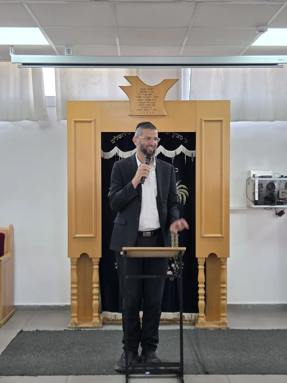 איש בחליפה מדבר במיקרופון לפני ארון הקודש - יום עיון בח׳ בתמוז