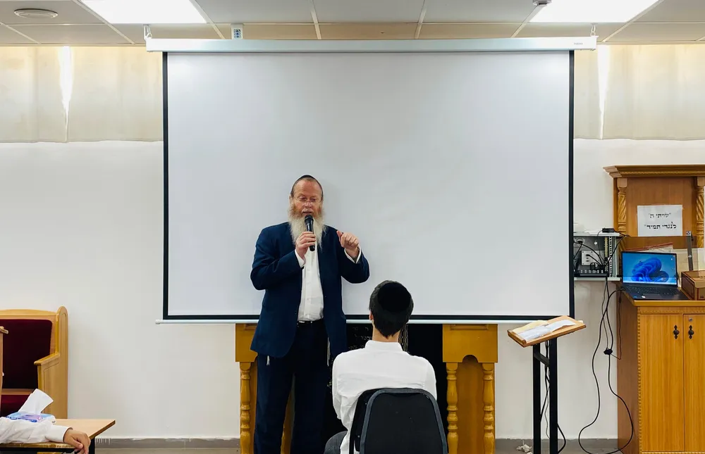 מרצה מזוקן מדבר במיקרופון מול מסך הקרנה - קולות מבחוץ
