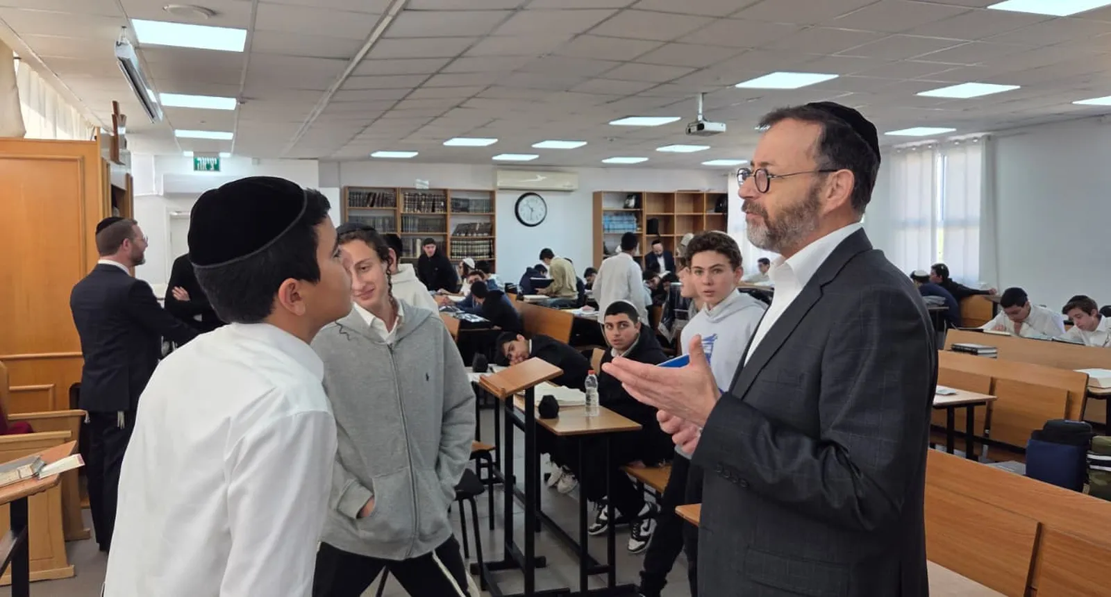 איש צוות בחליפה משוחח עם תלמידים בין שולחנות הלימוד - מבחן של חורף