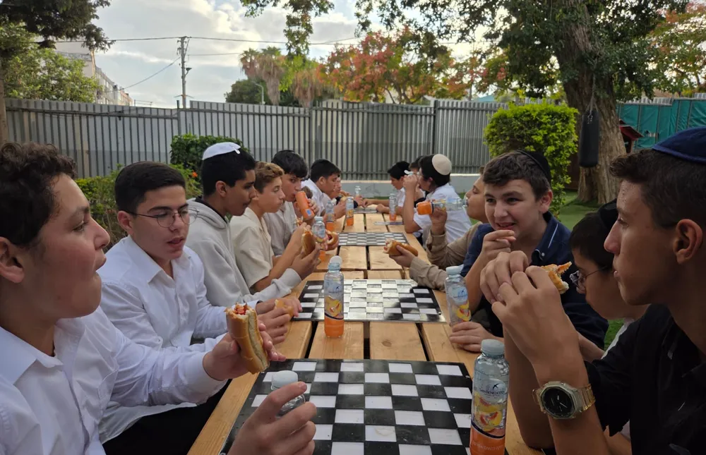 תלמידים אוכלים כריכים סביב שולחן בחצר - בזוגות מול הספר
