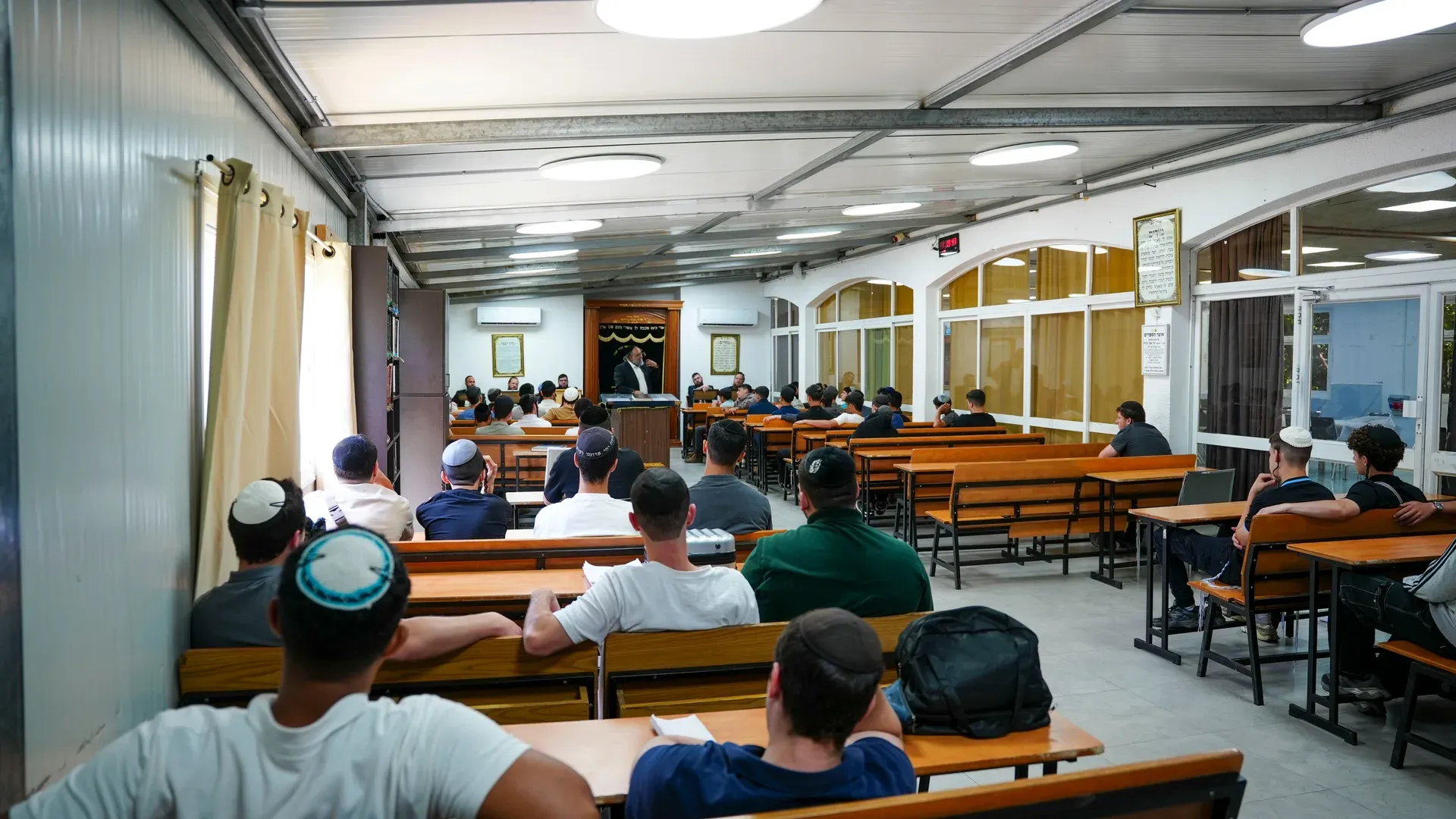 תלמידים יושבים בבית מדרש עם גג פח, בתאורה טבעית